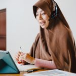 Asia muslim lady wear headphone using digital tablet talk to colleagues about sale report in conference video call while working from home at kitchen. Social distancing, quarantine for corona virus.